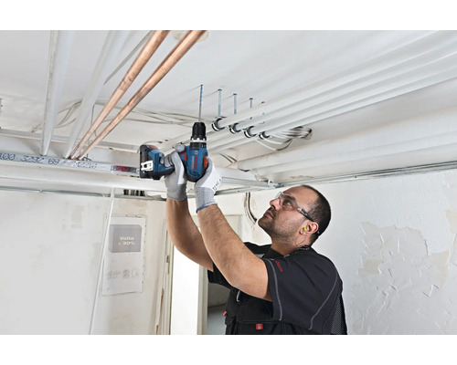 Vakman boort met Bosch accuschroefmachine in een betonnen plafond met leidingen op een bouwplaats.