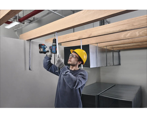 Vakman met veiligheidshelm en bril monteert een houten plafondconstructie met een accuboormachine op een bouwplaats.
