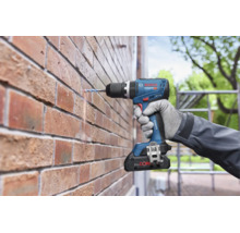 Een Bosch Professional accuboormachine boort met een boor in een bakstenen muur, vastgehouden door een hand met werkhandschoen.