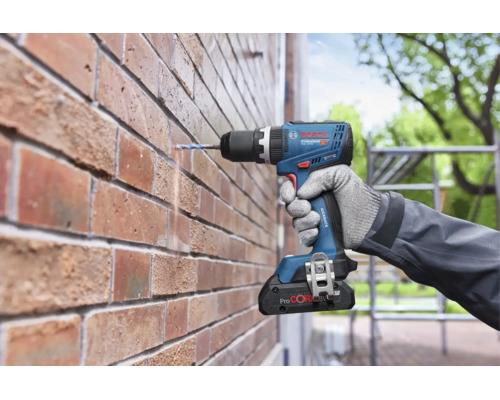 Een Bosch Professional accuboormachine boort met een boor in een bakstenen muur, vastgehouden door een hand met werkhandschoen.