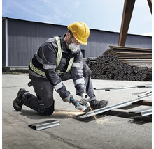 Vakman met veiligheidshelm en stofmasker snijdt een metalen profielrail met een haakse slijper op een bouwplaats.
