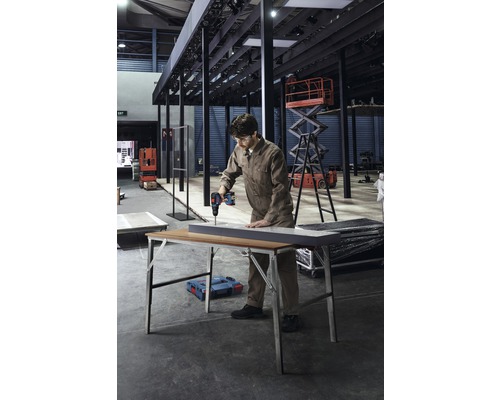 Ambachtsman boort een werkstuk op een werkbank met een accuschroefboormachine. Op de achtergrond zijn hefplatforms en andere werkplaatsapparatuur te zien.