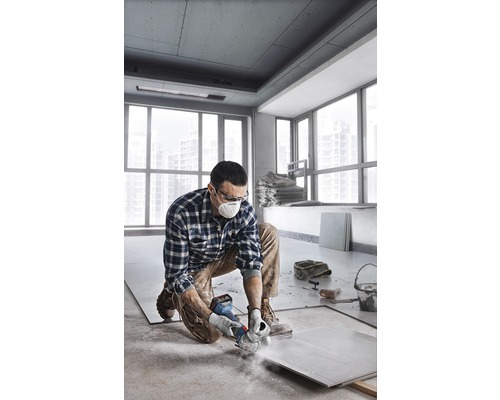 Een man knielt in een kamer en snijdt een tegel met een slijpmachine. Hij draagt een veiligheidsmasker en een veiligheidsbril.