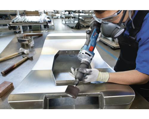 Een man bewerkt een metalen onderdeel met elektrisch gereedschap in een werkplaats. Er wordt persoonlijke beschermingsmiddelen gedragen.