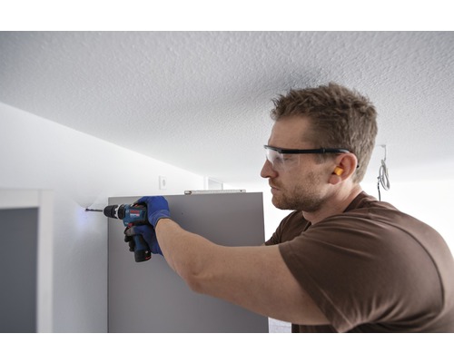 Een man boort met een boormachine een gat in een muur, terwijl hij een veiligheidsbril en handschoenen draagt.