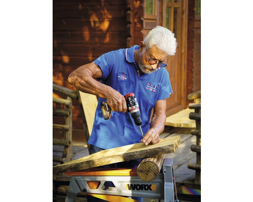 Een man schroeft een houten plank vast met een accuboormachine, die op een werkbank ligt.