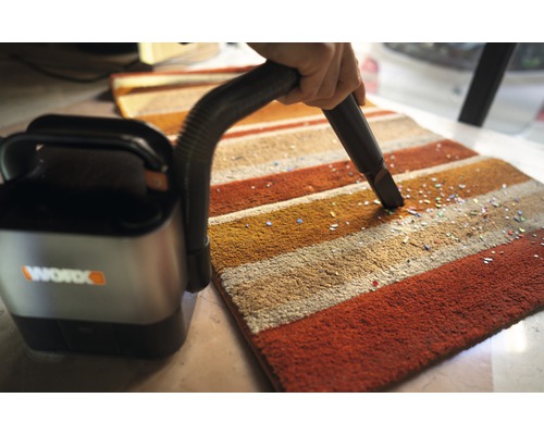 Een draadloze stofzuiger wordt gebruikt op een gestreept tapijt om kleine deeltjes te verwijderen. Worx bedrijfslogo.