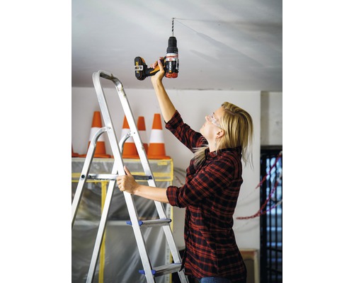 Vrouw boort met een boormachine een gat in het plafond terwijl ze op een ladder staat.