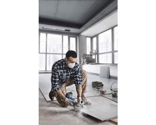 Een man knielt in een kamer en snijdt een tegel met een haakse slijper. Hij draagt beschermende kleding.
