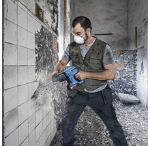 Man met veiligheidsbril en stofmasker werkt met een boorhamer aan een muur.