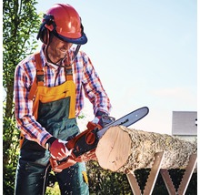 Man zaagt een blok hout met een elektrische kettingzaag op een zaagbok, terwijl hij een helm met vizier en gehoorbescherming draagt
