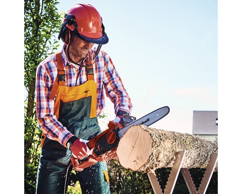 Man zaagt een blok hout met een elektrische kettingzaag op een zaagbok, terwijl hij een helm met vizier en gehoorbescherming draagt