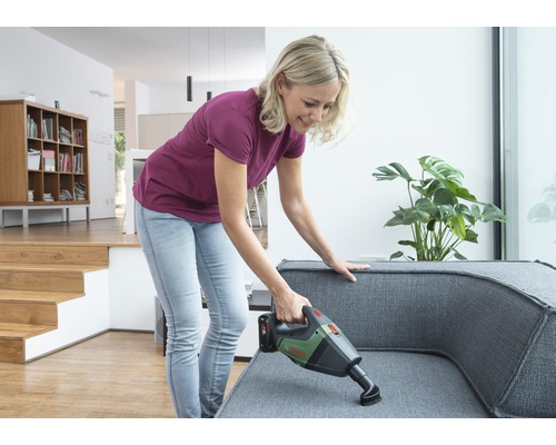 Vrouw stofzuigt met een accuzuiger een grijze bank in een lichte woonkamer.