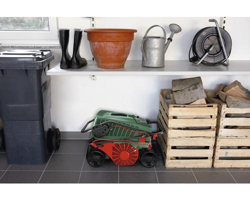 Foto van een opbergruimte met tuingereedschap, laarzen, vuilnisbak, grasmaaier, houten kisten en rekken.
