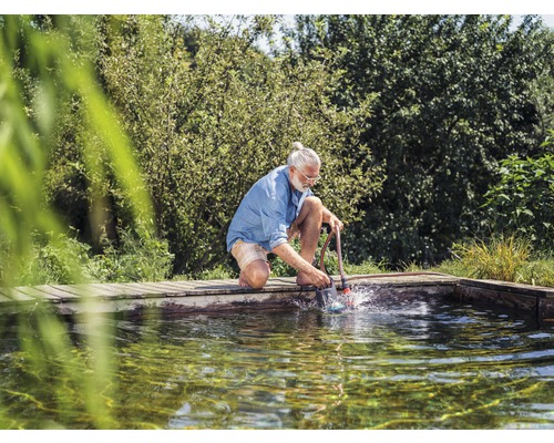Een man knielt aan de rand van de vijver en reinigt de vijver met een vijverslibzuiger.