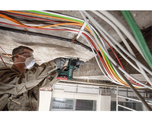 Een man boort met een boorhamer in een betonnen plafond, omgeven door kabels.