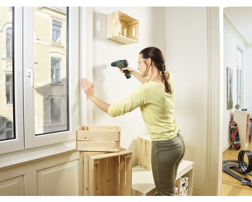 Vrouw boort houten kist met een boormachine aan de muur om een plank te maken