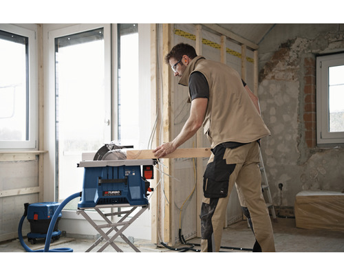 Een man gebruikt een tafelzaag om een houten plank te zagen. Een stofzuiger staat ernaast.