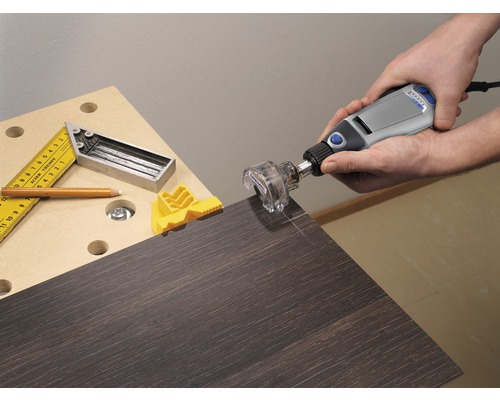 Een persoon gebruikt een Dremel multifunctioneel gereedschap met een freesopzetstuk op een donkere houten plaat. Een winkelhaak, een potlood en een gele werkstukspanner liggen op de werkbank.