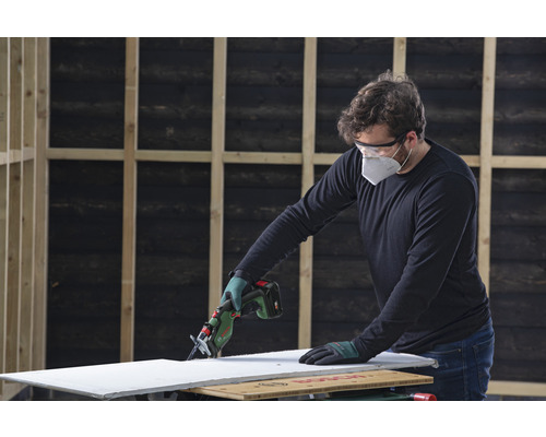 Een persoon zaagt een gipsplaat met een accuschrobzaag. Er wordt veiligheidsuitrusting gedragen.