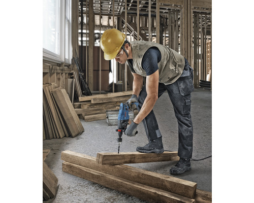 Een bouwvakker boort met een boormachine een gat in een houten plank op een bouwplaats.