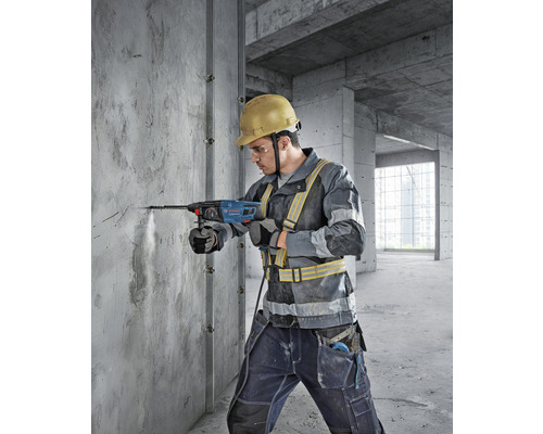 Een bouwvakker boort met een boorhamer in een betonnen muur. De man draagt een veiligheidshelm en veiligheidskleding.