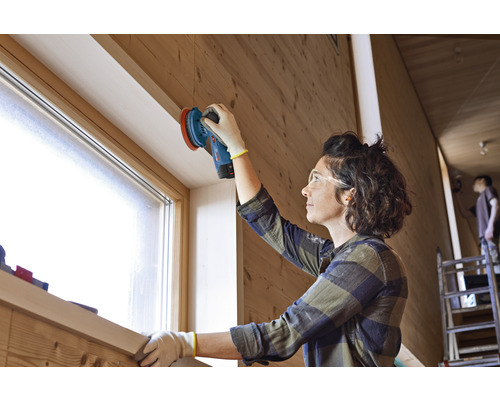 Vrouw schuurt hout met een schuurmachine in een lichte ruimte.