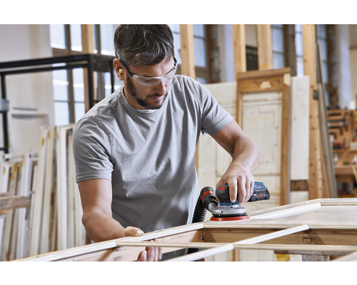 Een man schuurt een houten frame met een excentrische schuurmachine in een werkplaats. Hij draagt een veiligheidsbril en oordopjes.