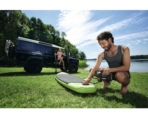 Man pompt een stand-up paddleboard op met een elektrische pomp op het gazon, met een terreinwagen en een vrouw op de achtergrond