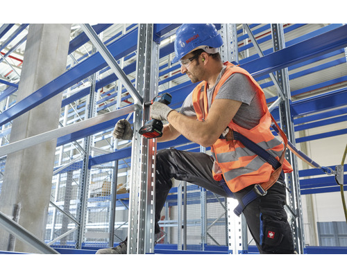 Man monteert magazijnstelling met accuschroevendraaier, veiligheidshelm en veiligheidsvest in een magazijn