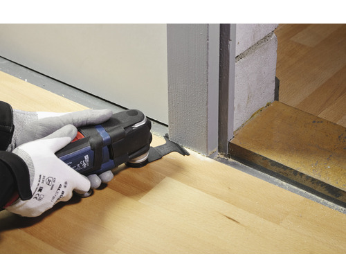 Een persoon gebruikt een multifunctioneel gereedschap om een houten vloer bij een deur te zagen.