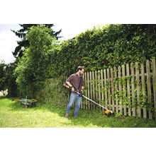 Een man trimt gras met een accugrastrimmer in een tuin met een houten hek en een heg.