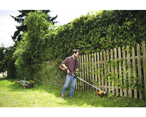 Een man trimt gras met een accugrastrimmer in een tuin met een houten hek en een heg.