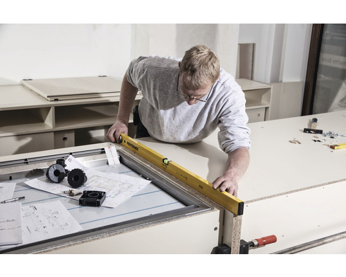 Een man gebruikt een waterpas op een werkbank. Bouwtekeningen en gereedschap liggen klaar.