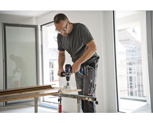 Een man boort een gat in een houten plank met een accuboormachine. Hij draagt een veiligheidsbril en werkkleding.