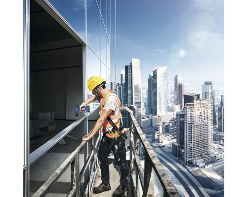 Een bouwvakker met veiligheidshelm en veiligheidsvest gebruikt een accuschroevendraaier op een steiger met wolkenkrabbers op de achtergrond.