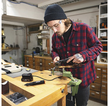 Man werkt met een haakse slijper aan een werkstuk dat is vastgeklemd in een bankschroef op een werkbank. Ook te zien: een multifunctioneel gereedschap en een accu.