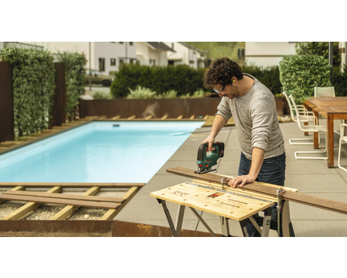Man zaagt houten plank met een decoupeerzaag in de tuin, op de achtergrond een zwembad