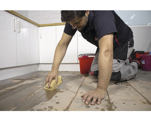 Een man maakt tegels schoon met een spons in een ruimte met keukenkasten.