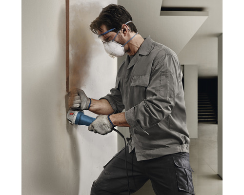 Ambachtsman schuurt een muur met een haakse slijper, terwijl hij een veiligheidsbril en een stofmasker draagt.