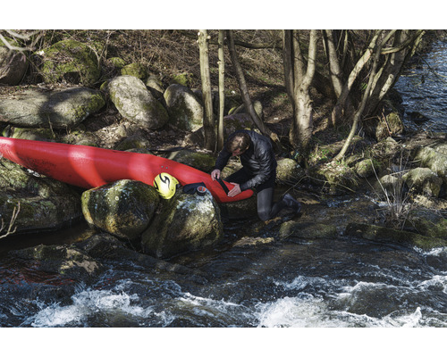 Man repareert kajak aan rivieroever met rotsen en bomen.