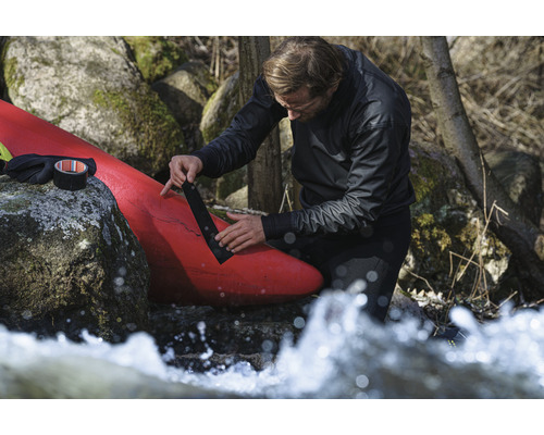 Een man repareert een rode kajak met zwarte ducttape in de buurt van rotsen en stromend water.