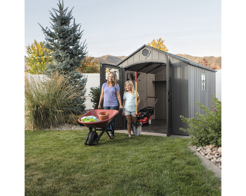 Scene met een moeder, een kind, een tuinhuis, een kruiwagen en tuingereedschap in een tuin.
