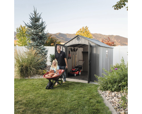 Scene met een gereedschapsschuur waarin een grasmaaier en tuingereedschap zijn opgeslagen, terwijl een man een meisje in een kruiwagen duwt.