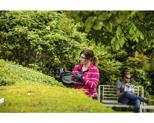 Een vrouw snoeit een haag met een heggenschaar in de tuin, terwijl een man op een bank zit te ontspannen.