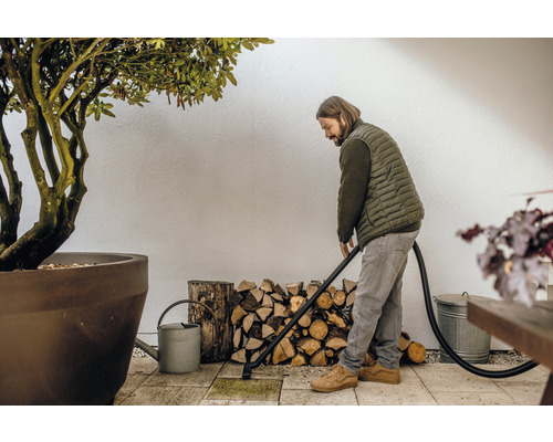 Een man reinigt een terras met een aszuiger, naast hem staan een boom, brandhout en een gieter