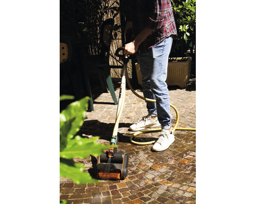 Een man maakt bestrating schoon met een borstelreiniger en waterslang in de tuin.