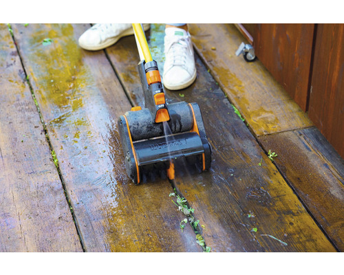 Een persoon reinigt een houten terras met een borstel en water.