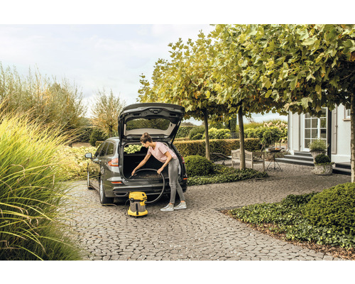Een vrouw maakt de kofferbak van een auto schoon met een gele stofzuiger van Kärcher, op de achtergrond een huis met een tuin