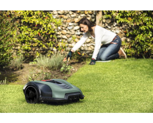 Een robotmaaier staat op een gazon, terwijl een vrouw op de achtergrond in de tuin werkt.